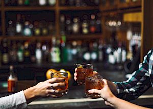 Three people clink glasses filled with beverages at a bar, with shelves of bottles in the background.