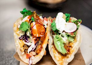 The image shows two loaded tacos with various toppings including cilantro, radishes, and crema on a white plate with a brown napkin underneath.